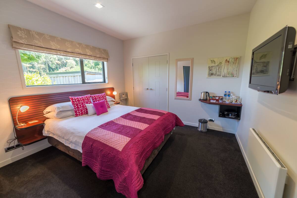 A bedroom with a bed, bedside table, lamp, window with blinds, door, and a small shelf with items.