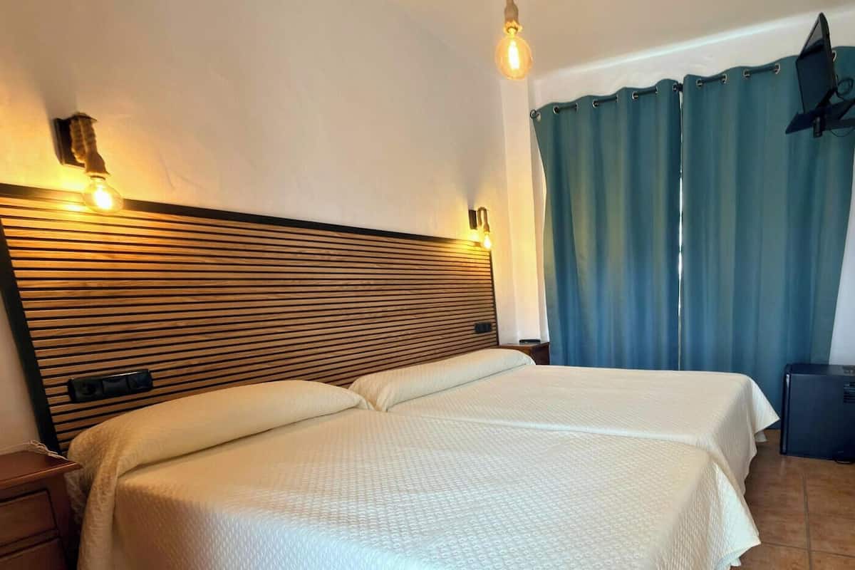 A double bed with a wooden headboard, a bedside table, a wall-mounted light, and a blue curtain.