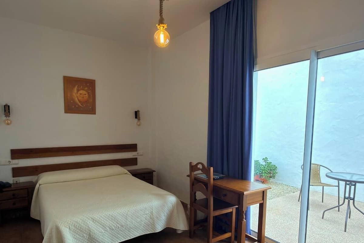 A hotel room with a bed, a desk, a chair, a table, a lamp, a window with curtains, and a view of the sea.
