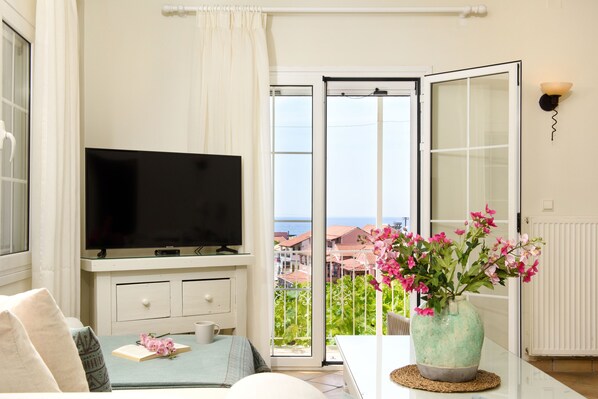 A living room with a television, a dining table with a vase of flowers, and a view of the sea through a sliding glass door.