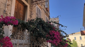 Un edificio con balcón de hierro forjado, puerta de madera y enredaderas de buganvilla rosadas.