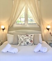 A neatly made bed with floral pillows, a window with curtains, and wall-mounted lamps.