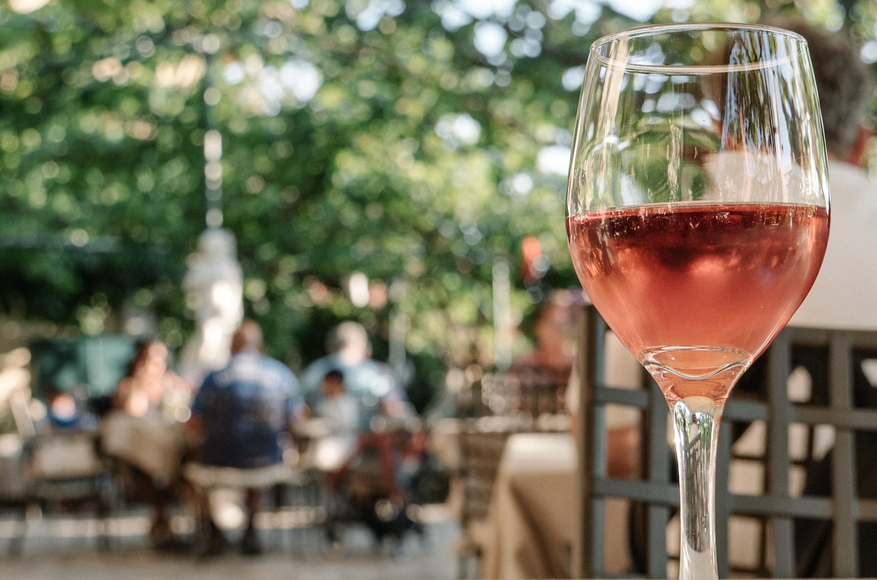 Una copa de vino tinto en una copa de martini, con un fondo desenfocado de personas y vegetación al aire libre.