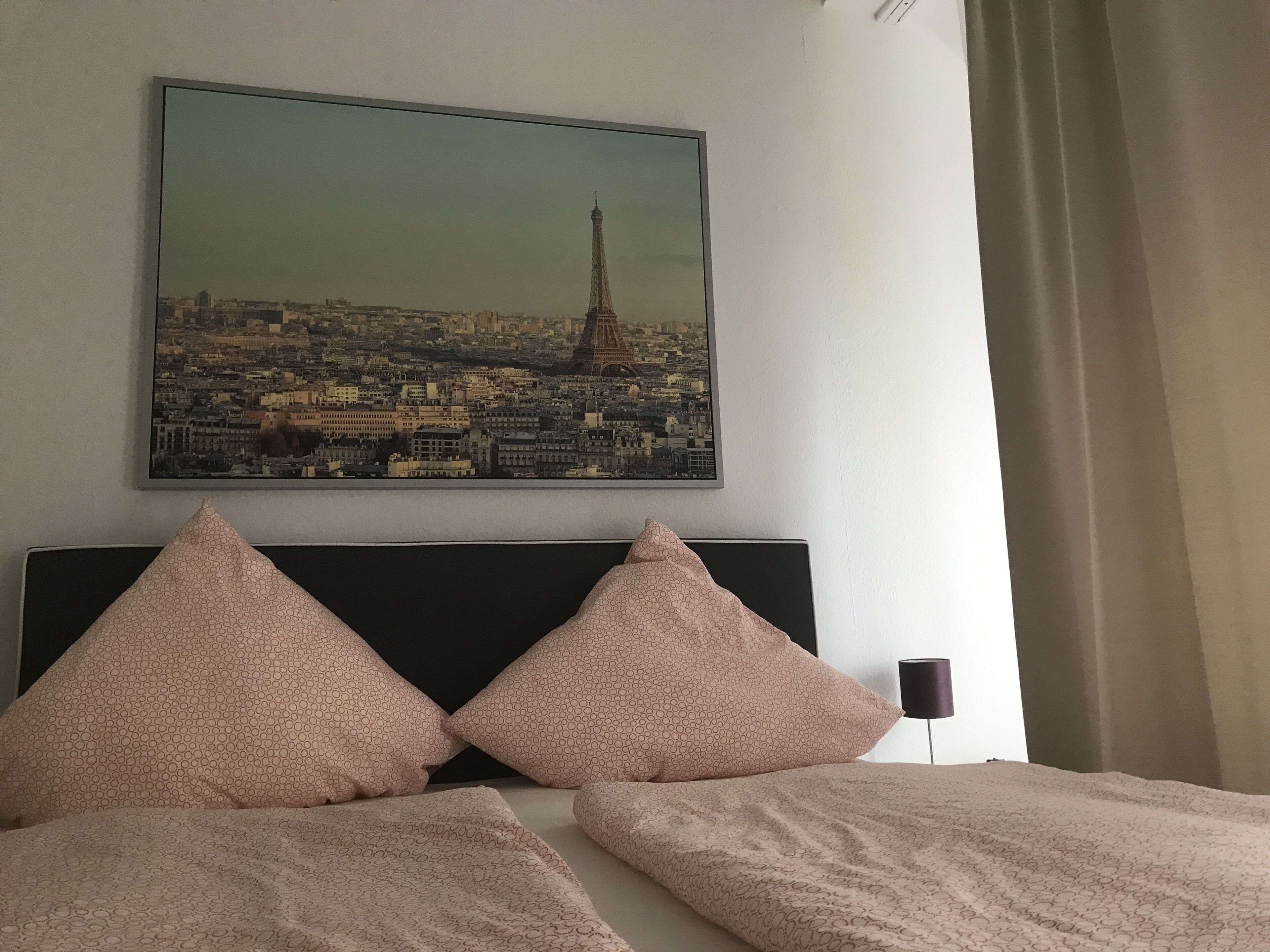 Une chambre d’hôtel avec un lit agrémenté de deux oreillers assortis, une tête de lit et un tableau représentant un paysage urbain, encadré et placé au-dessus du lit.