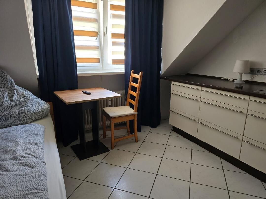 A small, compact room with a kitchenette, a dining area with a table and chair, and a window with blinds.