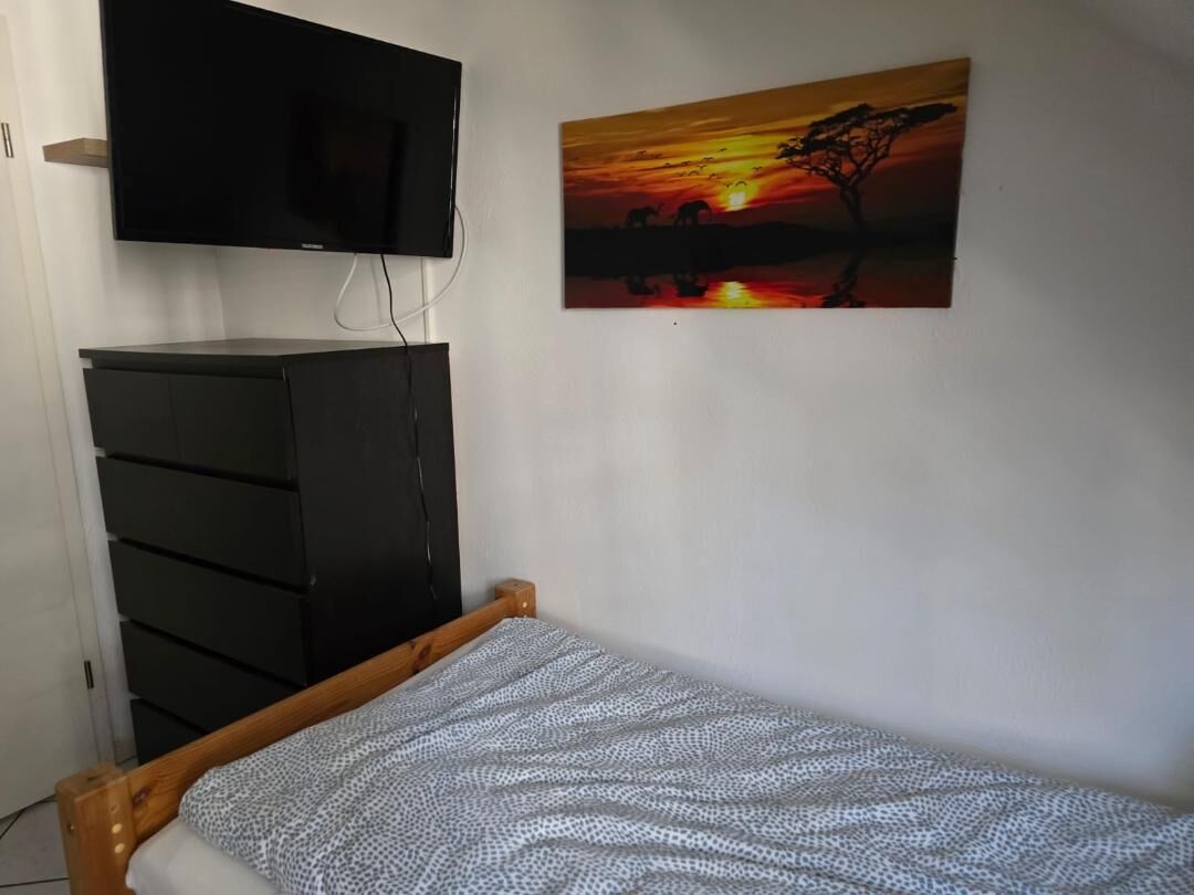 A bedroom with a bed, a TV mounted on the wall, a black wardrobe, and a sunset painting.