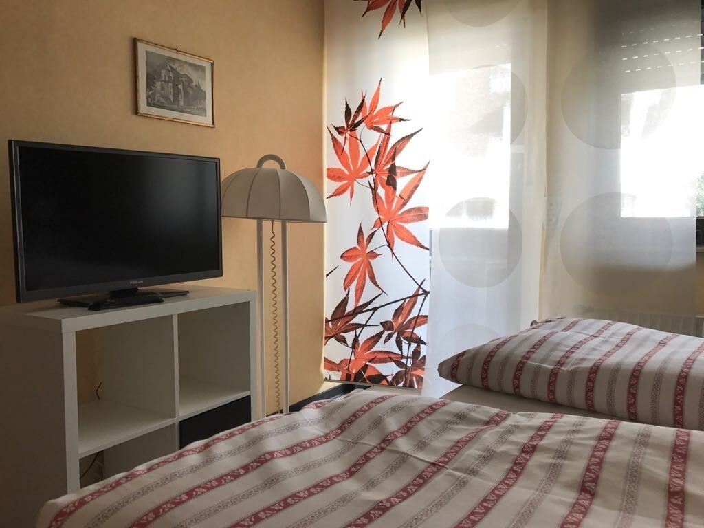 Une chambre d’hôtel avec un téléviseur posé sur un meuble blanc, une lampe, un lit avec une literie à rayures et un mur orné d’un motif de feuilles.