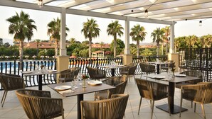 Un área de comedor al aire libre con mesas y sillas, con vista a una piscina y palmeras.