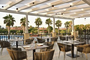 Un área de comedor al aire libre con mesas y sillas, con vista a una piscina y palmeras.