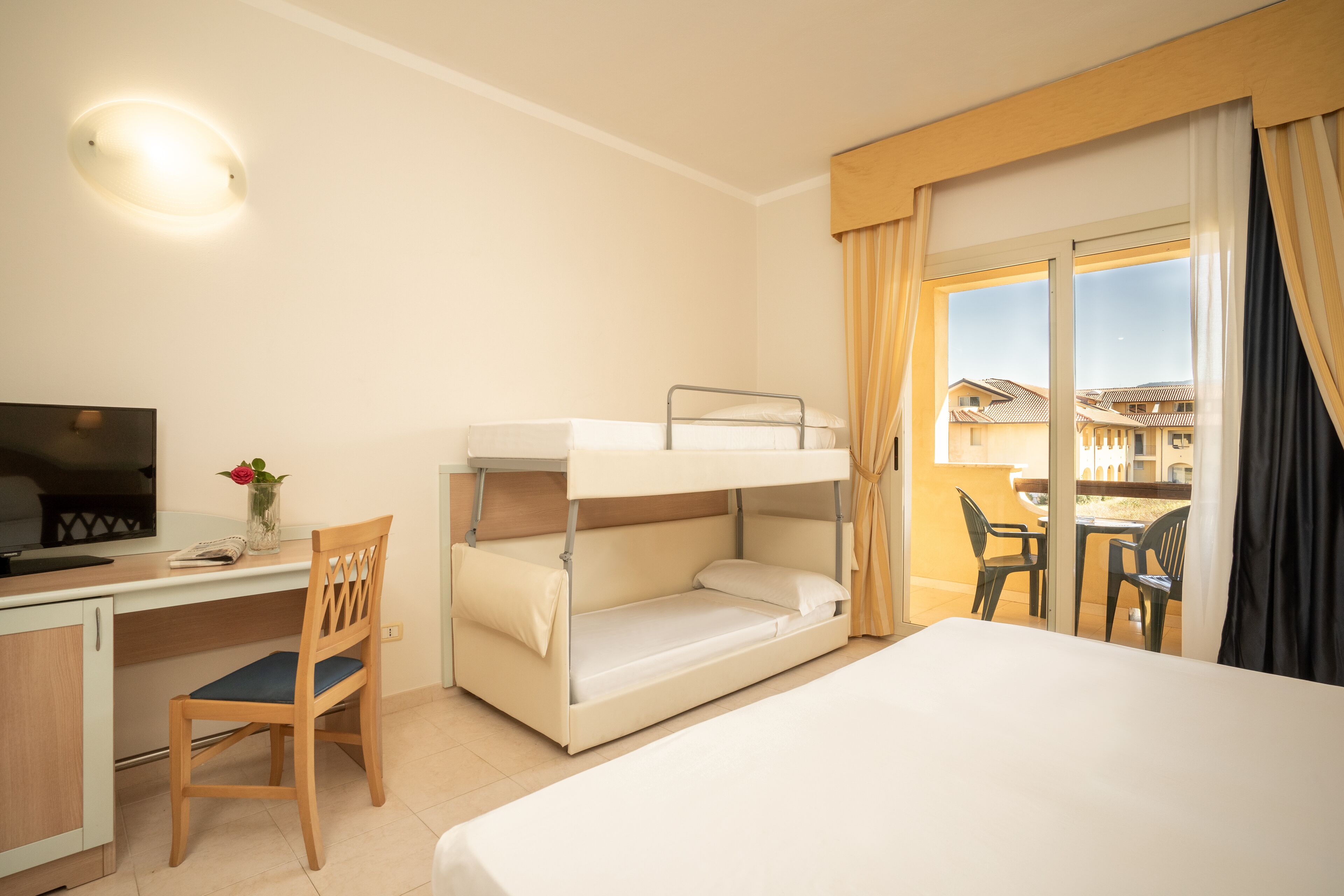 A hotel room with a bunk bed, a desk with a chair, a TV, and a view of buildings through a window.