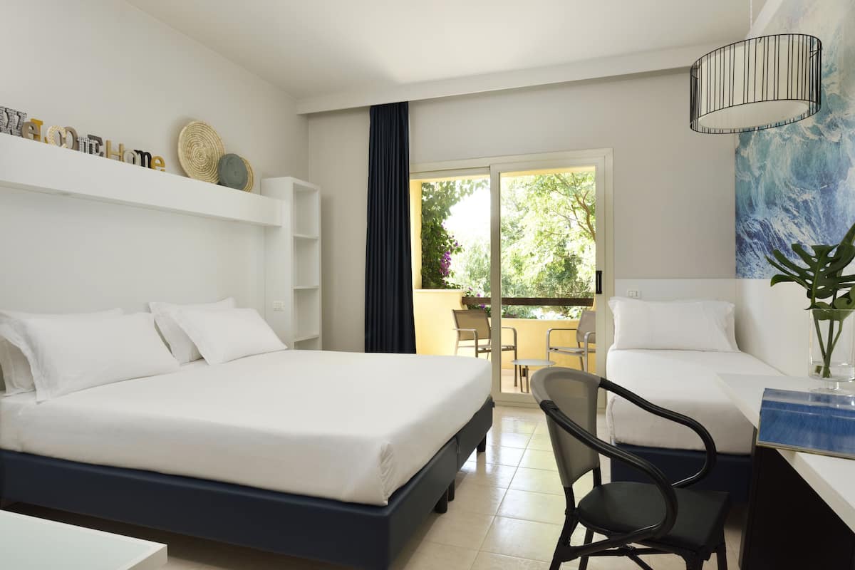 A hotel room with two beds, a desk, a chair, and a balcony with a view of greenery.
