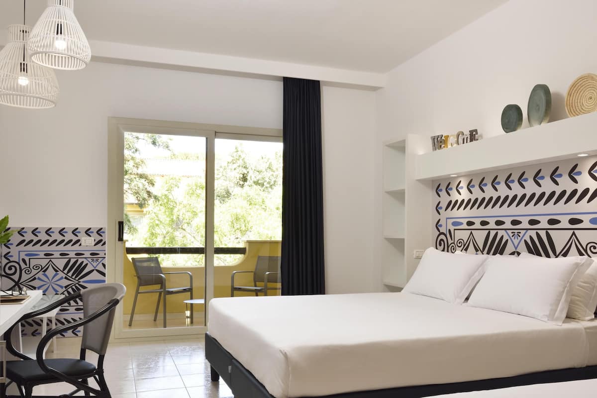 A hotel room with a bed, a desk with a chair, a balcony with a chair, and decorative wall art.