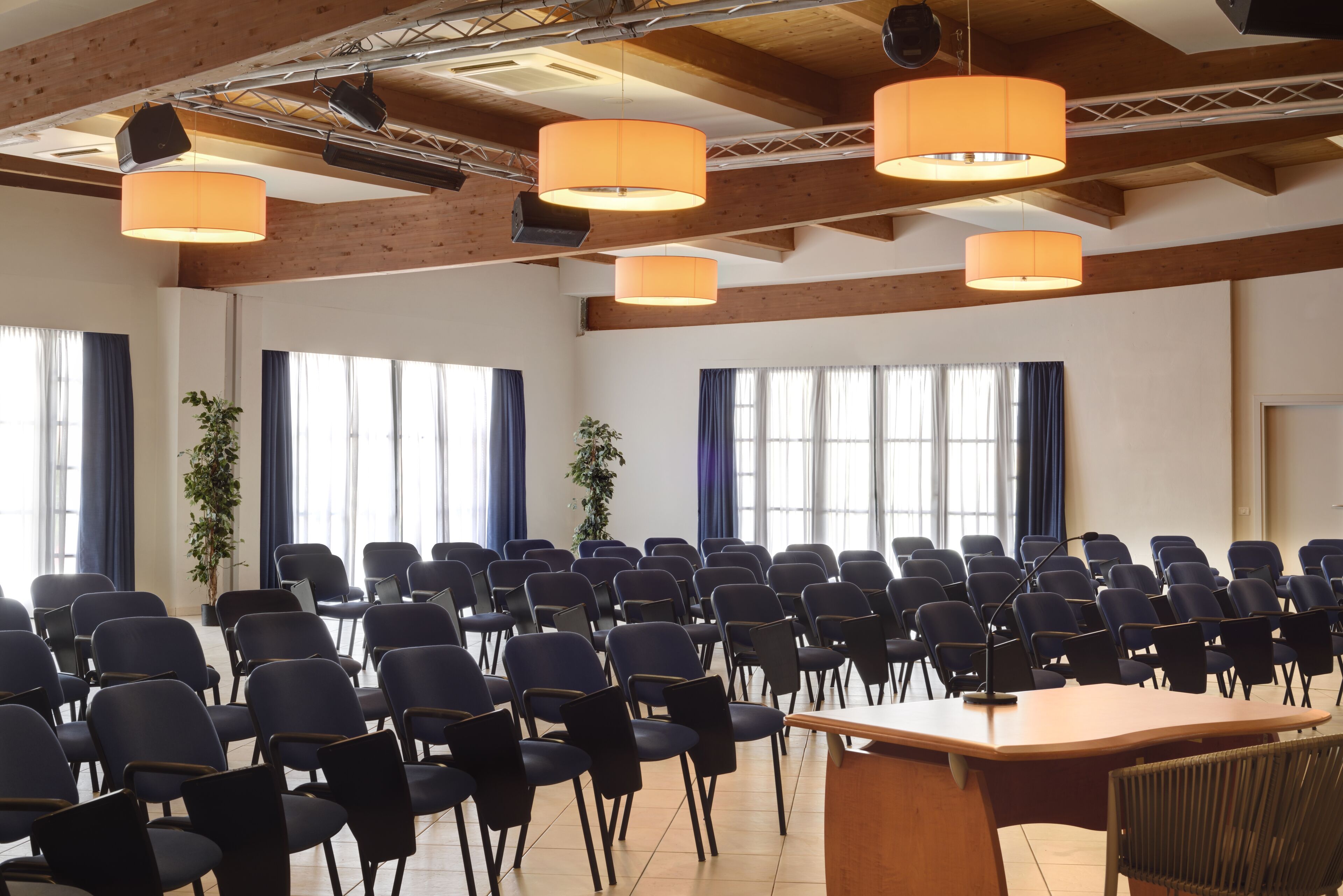 Ein Konferenzraum mit Reihen von Stühlen, einem großen Tisch und hängenden, orangefarbenen Leuchten.
