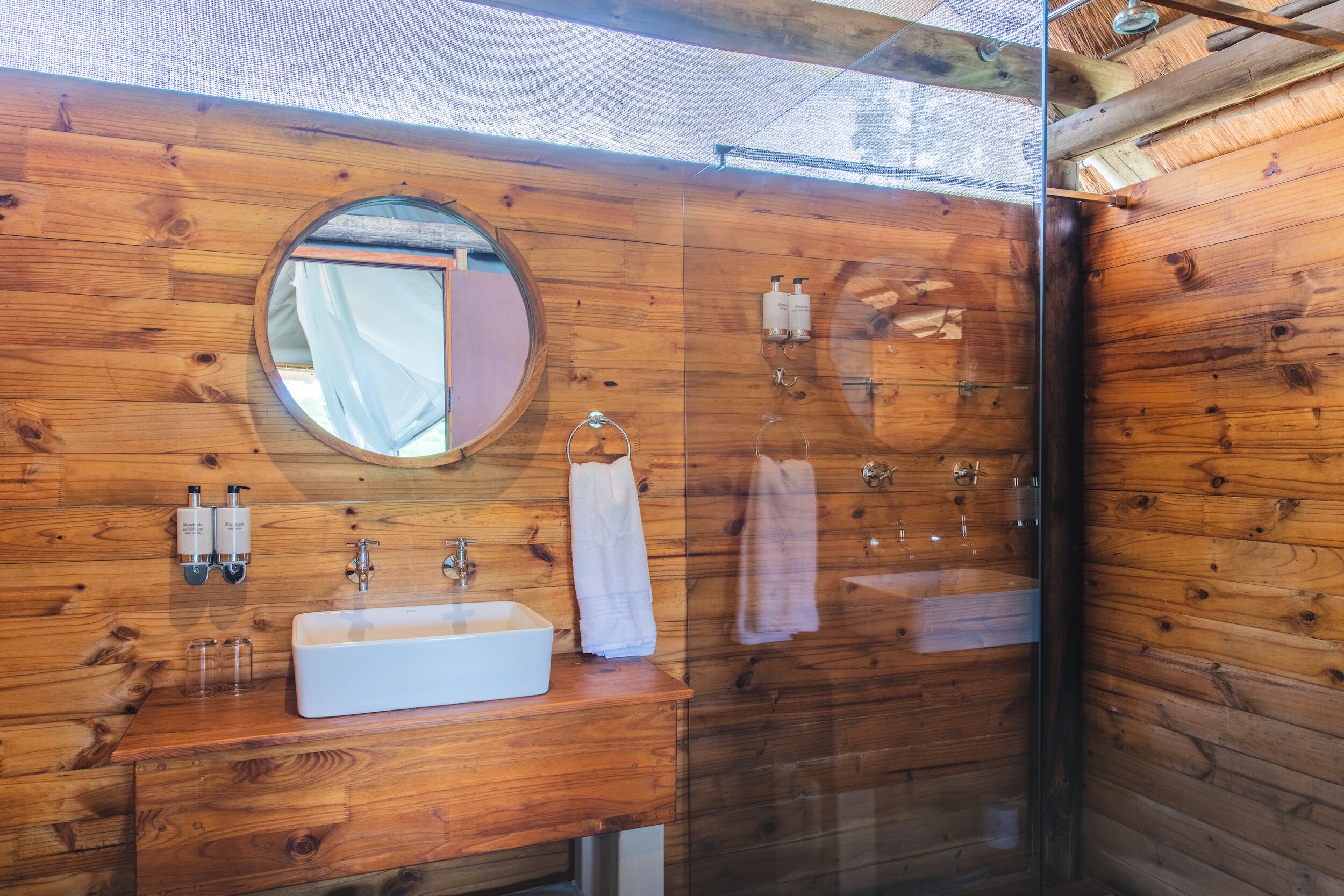 Un baño de madera con un espejo redondo, un lavamanos blanco y dos botellas de champú.