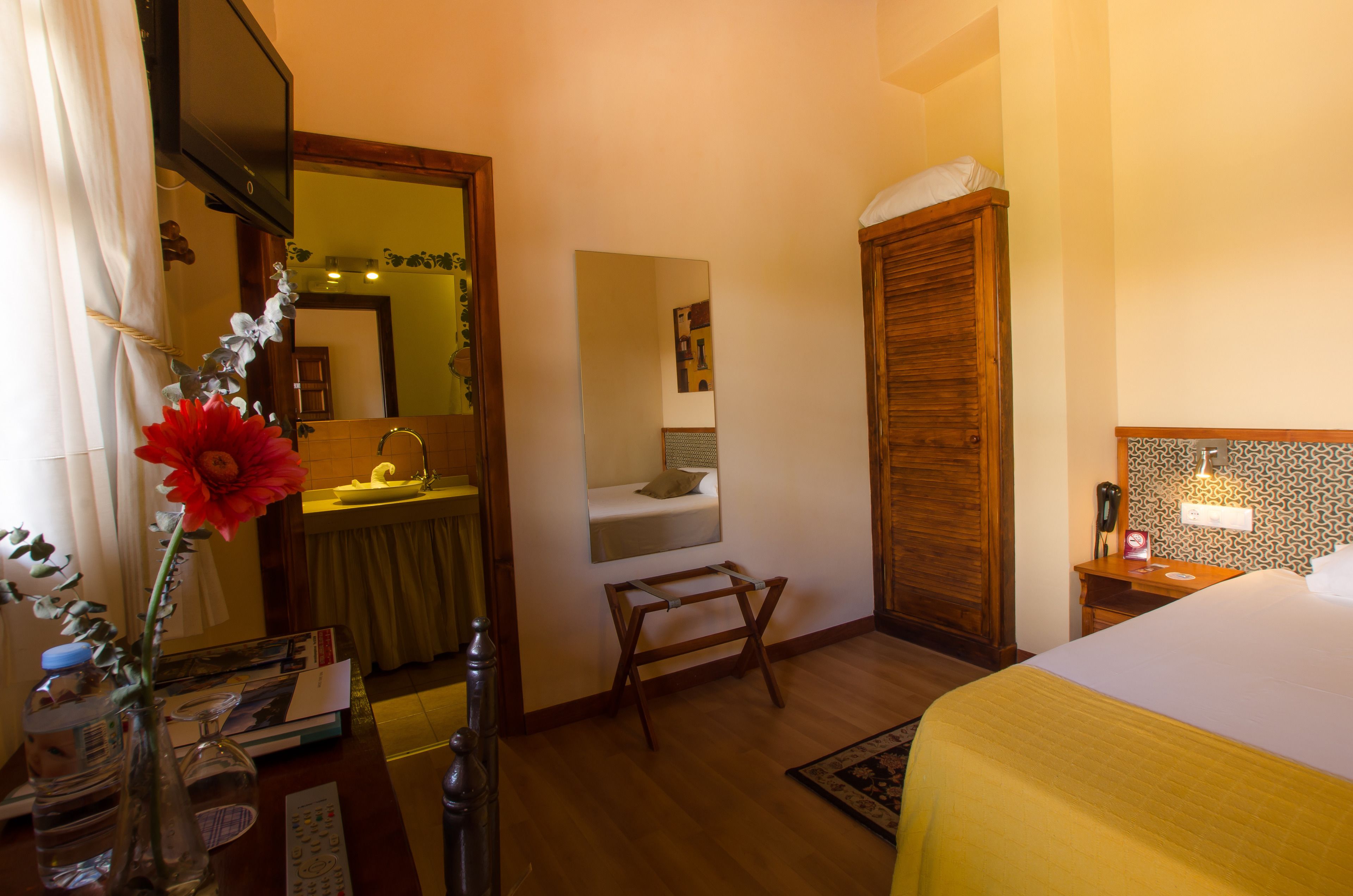 Habitación de hotel con cama, armario de madera, pequeña mesa de madera, espejo, lavabo y un jarrón con flores.