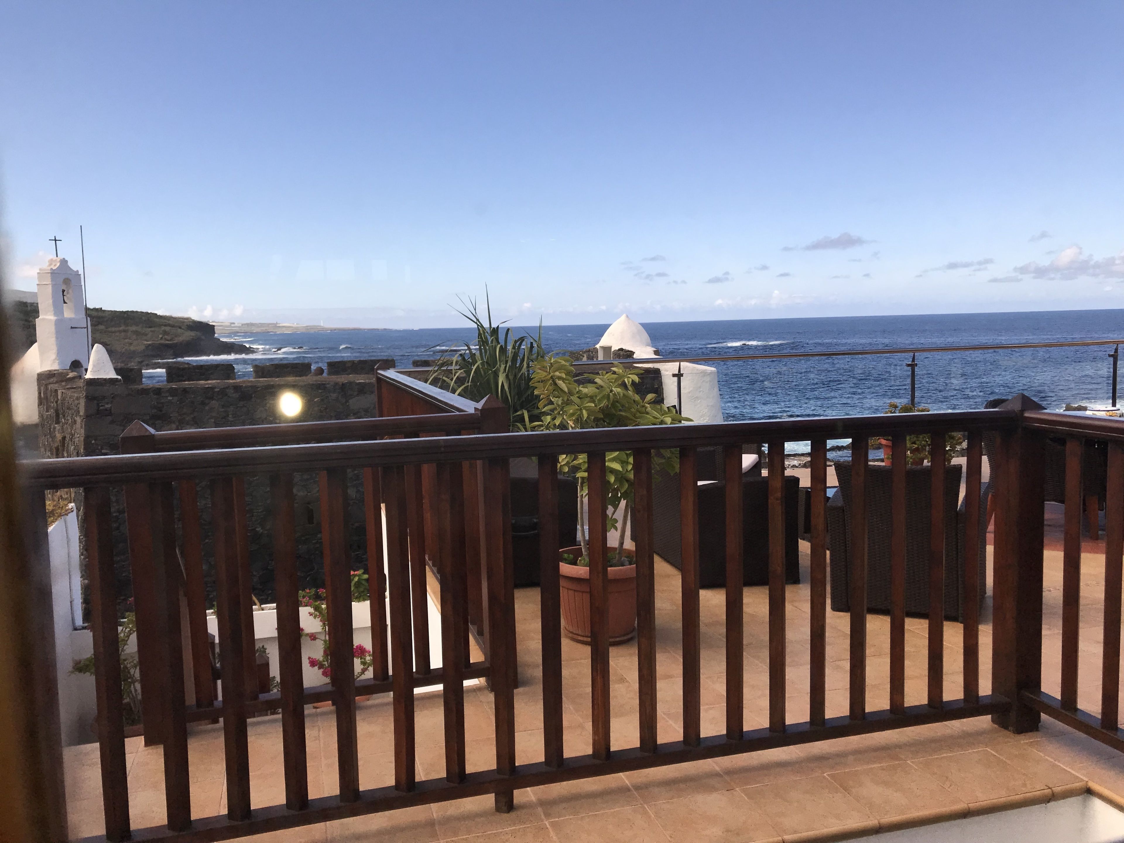 A balcony with a view of the sea, a white building with a cross, and potted plants.