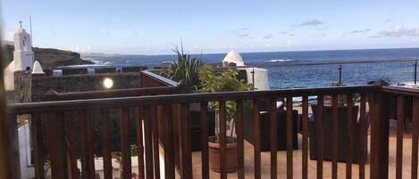 A balcony with a view of the sea, a white building with a cross, and potted plants.