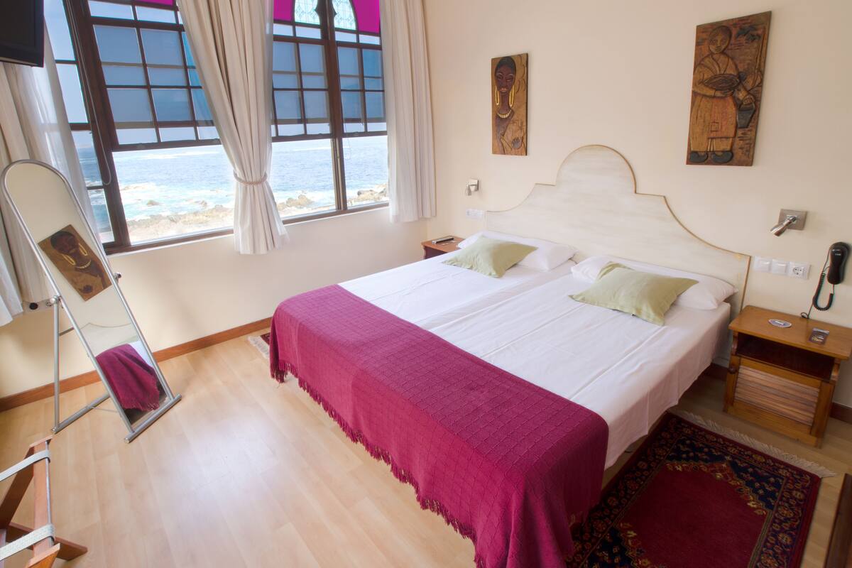 Una habitación de hotel con una cama grande, una mesita de noche, un espejo y vistas al océano.