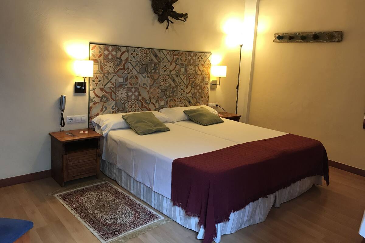 Una cama bien tendida con un cabecero estampado, dos lámparas de noche, una mesita de noche de madera y un adorno en la pared.