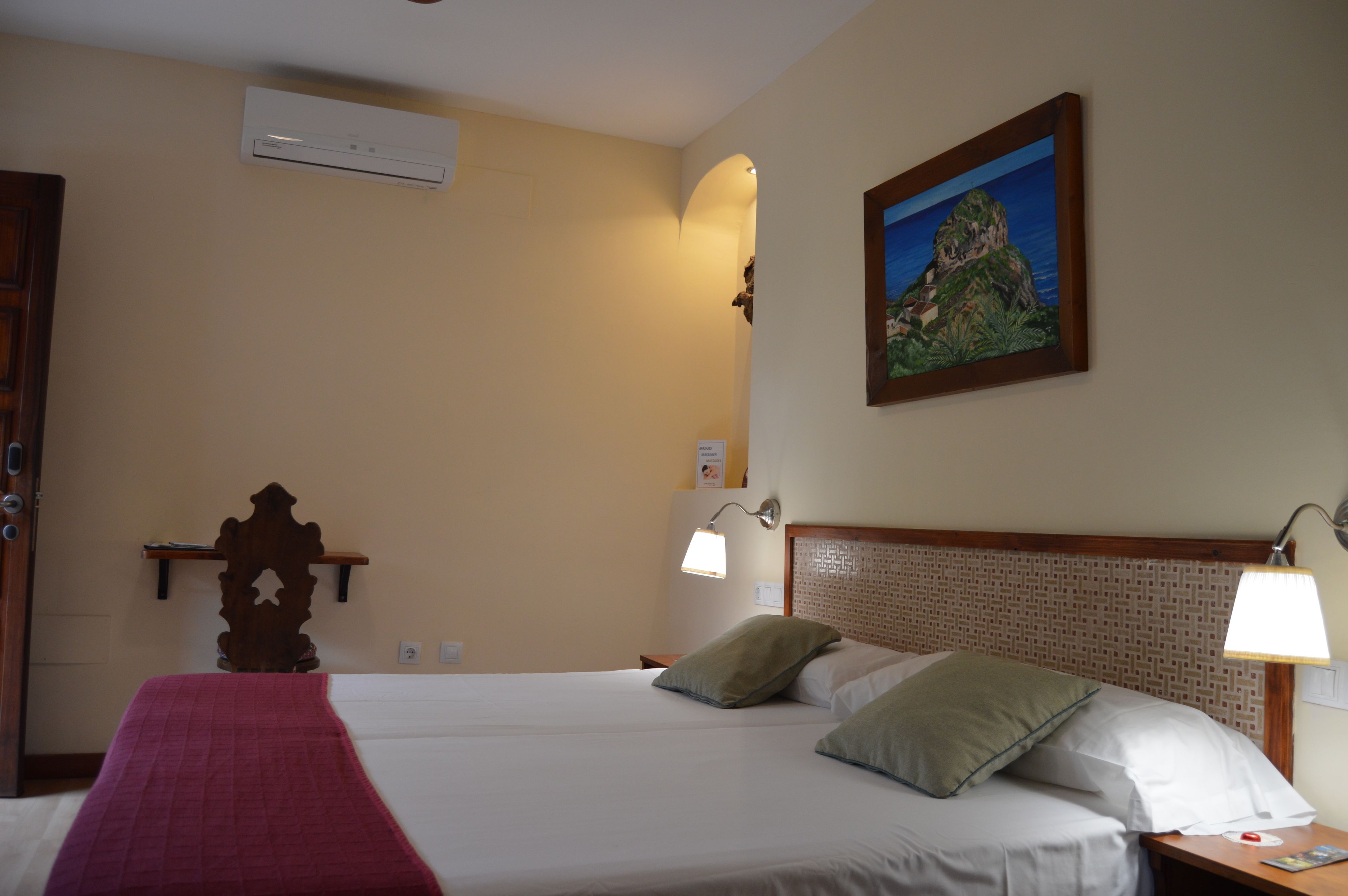 Habitación de hotel con cama, dos lámparas de noche, un cuadro en la pared, un equipo de aire acondicionado y un cabecero de madera.