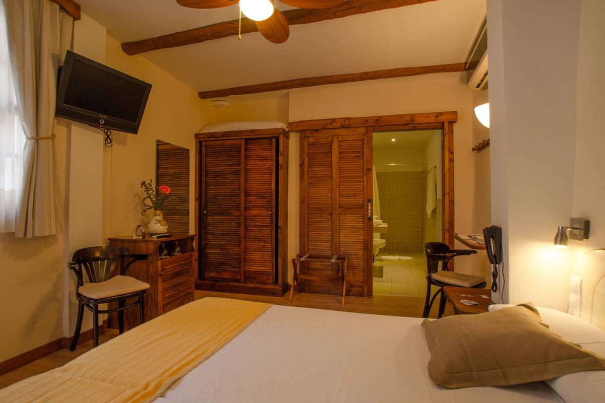 Un dormitorio con cama, armario de madera, escritorio con silla, televisor y baño visible a través de un vidrio.