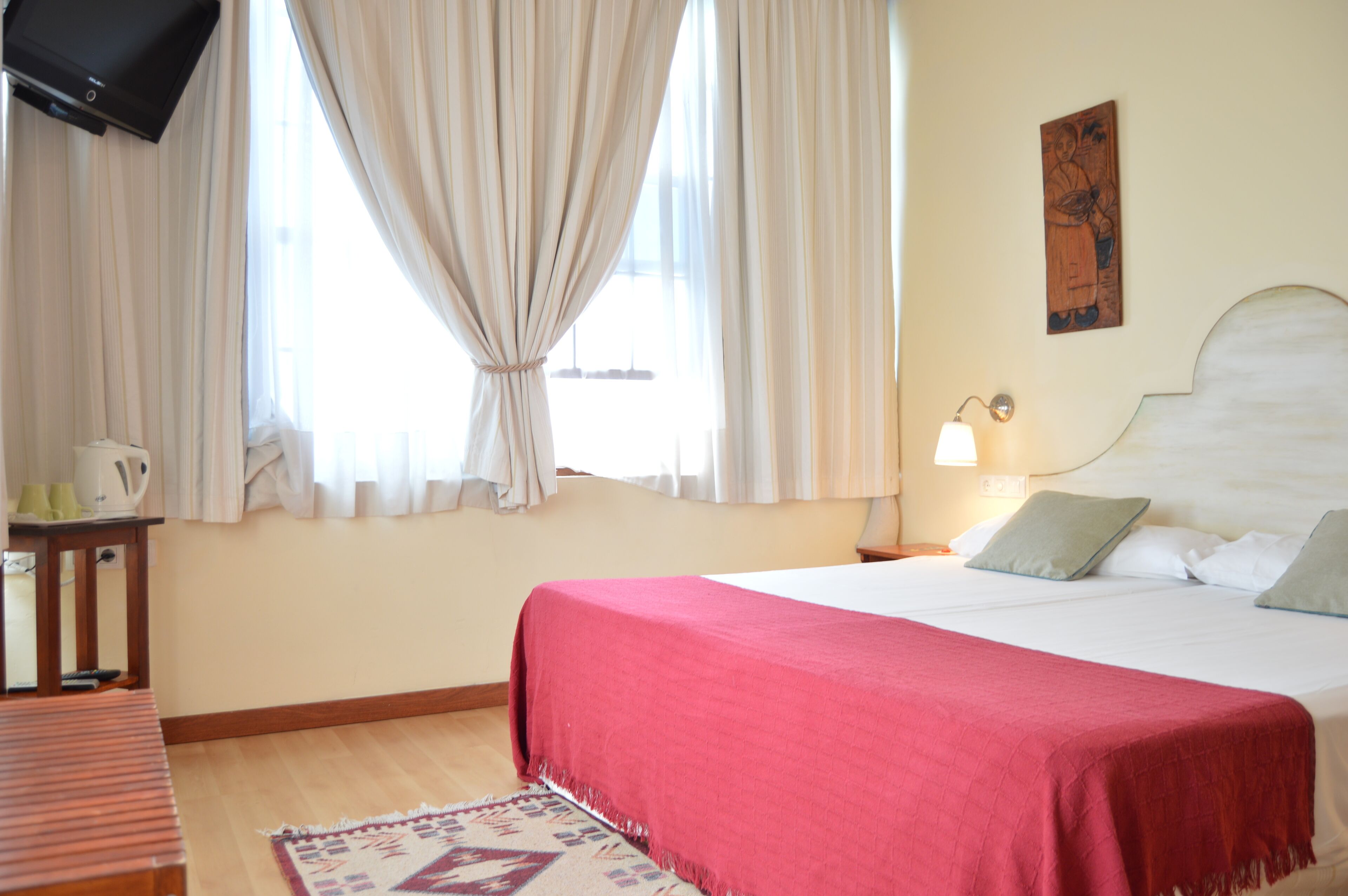 A hotel room with a bed, a television mounted on the wall, a bedside table with a lamp, and a window with curtains.