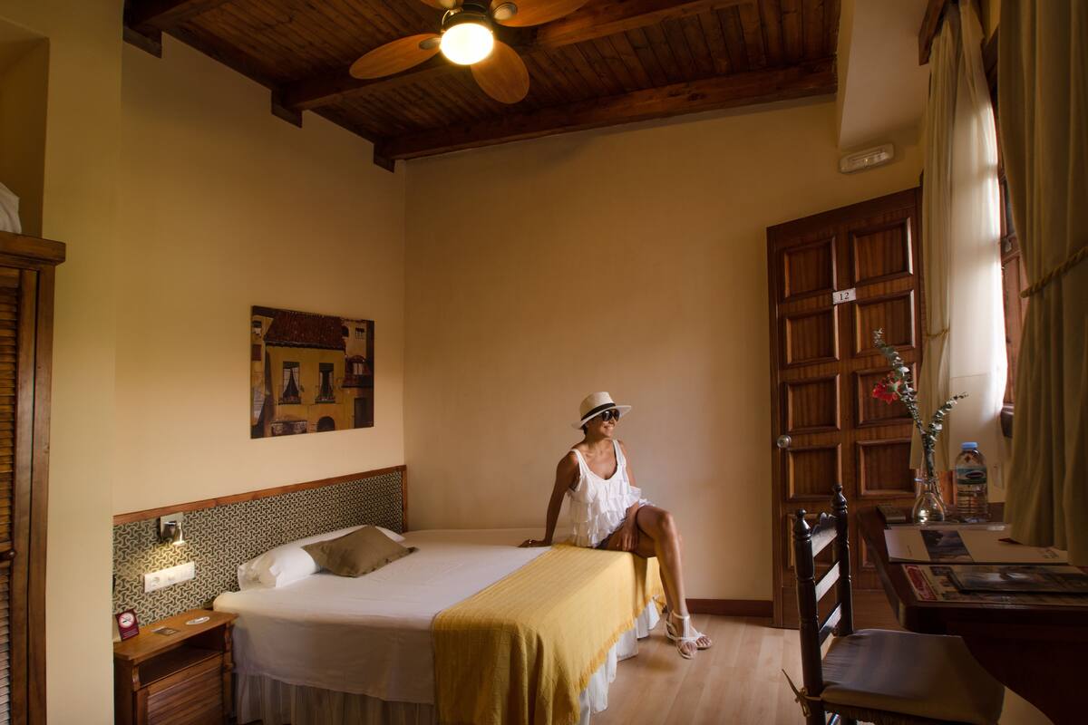 Una habitación con una cama, techo de madera, un escritorio, una silla, una mesita de noche, un reloj de pared, un cuadro enmarcado y una ventana con cortinas.