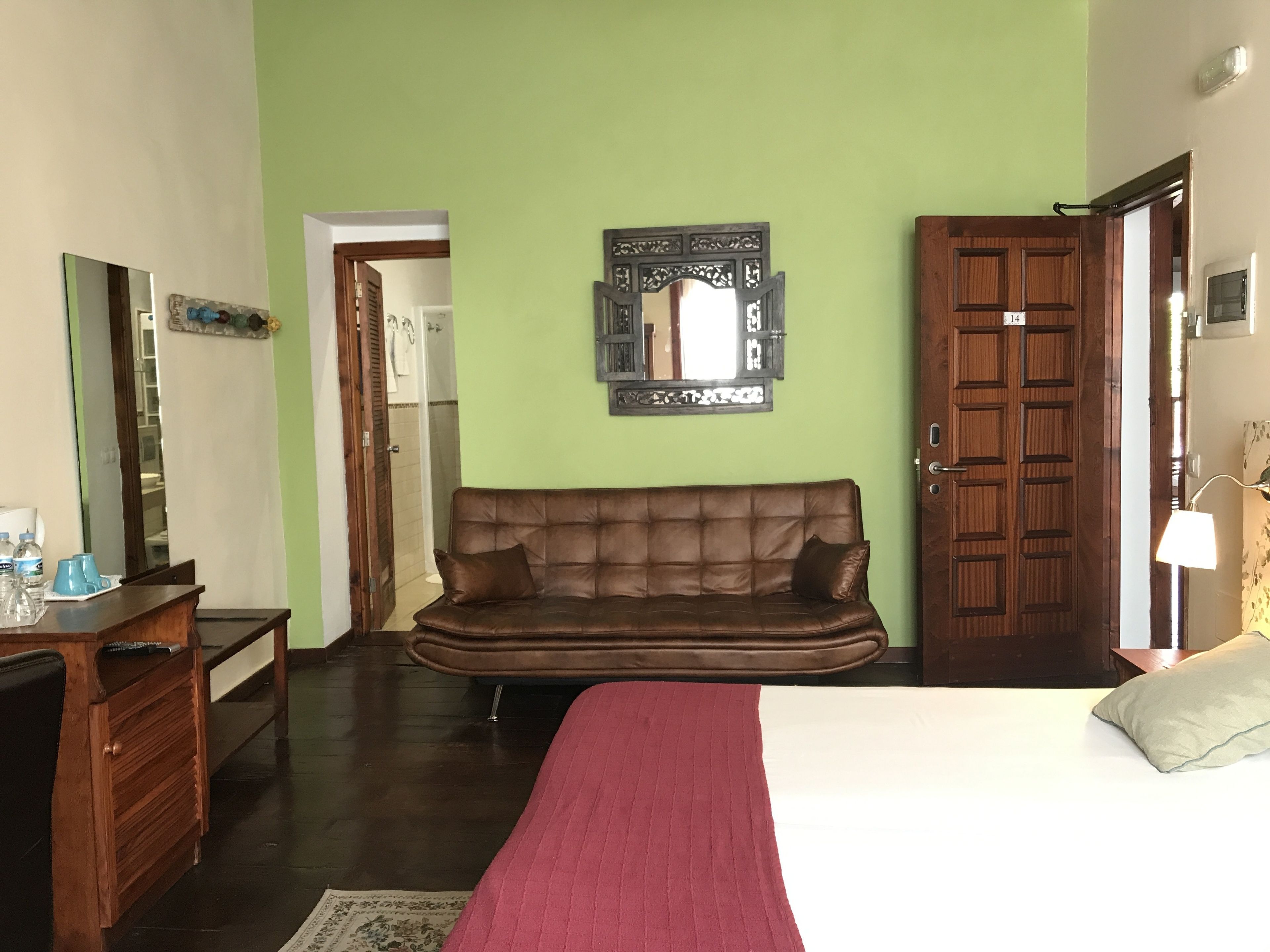 Une chambre avec un canapé en cuir marron, un lit recouvert d’une couverture rouge, une commode en bois, un miroir et une porte portant un numéro.