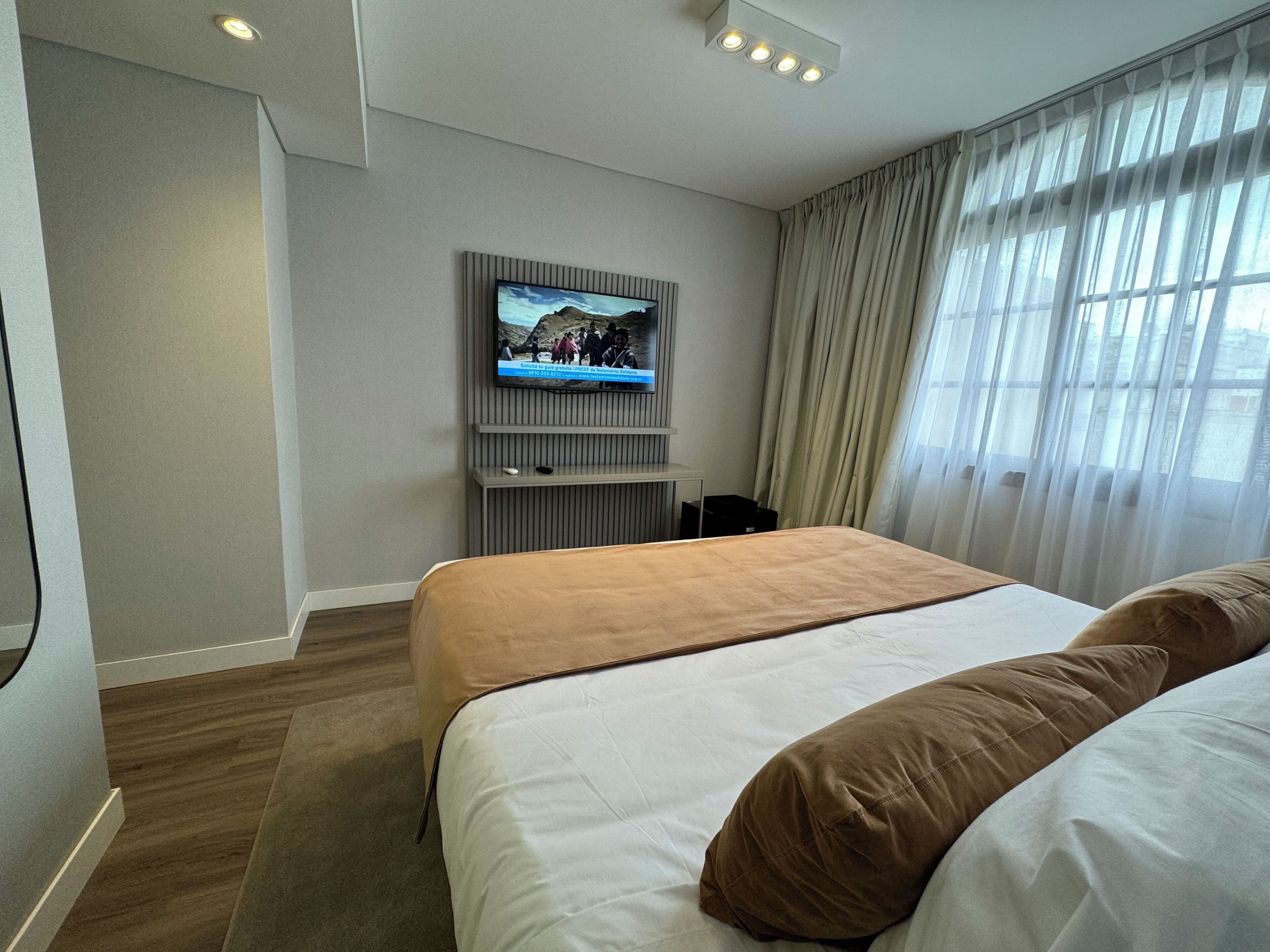 A hotel room with a bed, a television mounted on the wall, a window with curtains, and a wooden floor.