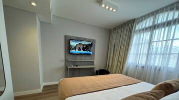 A hotel room with a bed, a television mounted on the wall, a window with curtains, and a wooden floor.