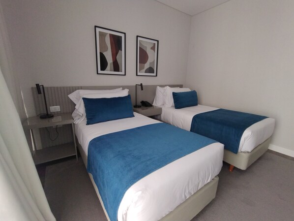 A hotel room with two beds, each with blue and white bedding, a desk with a lamp, and two abstract paintings on the wall. - Guemes Hotel & Spa (Salta)