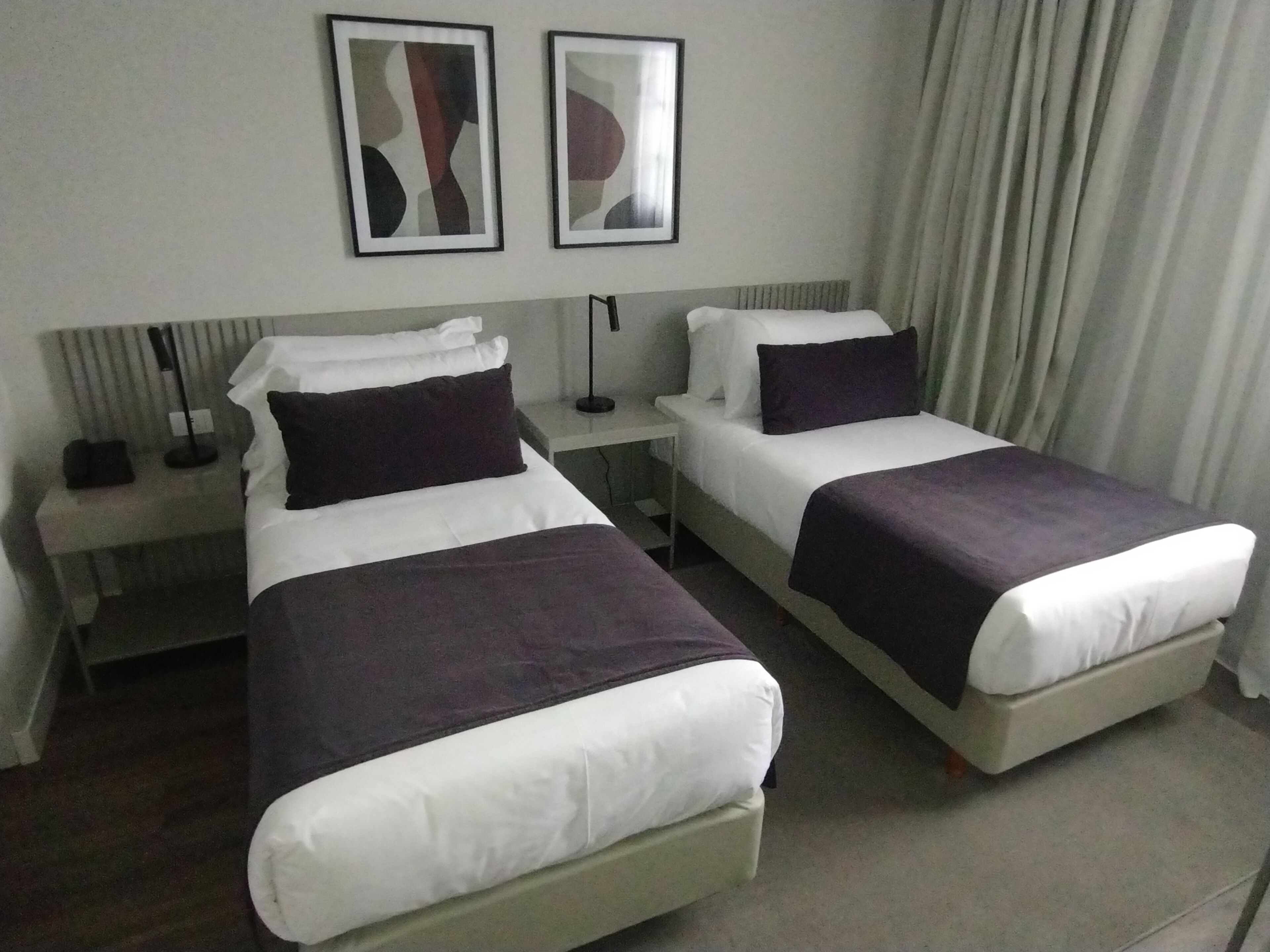 A hotel room with two single beds, a nightstand with a lamp, and two framed artworks on the wall.