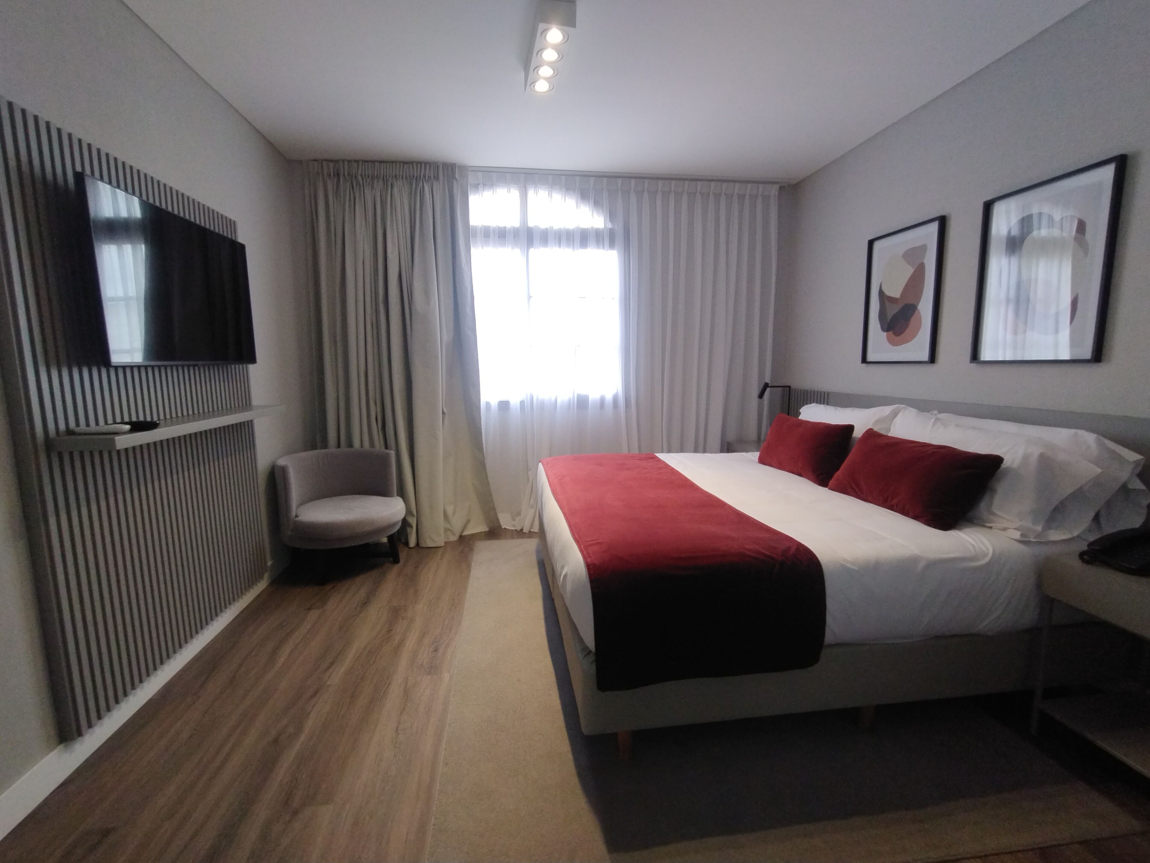 A hotel room with a bed, a chair, a TV mounted on the wall, and a window with curtains.
