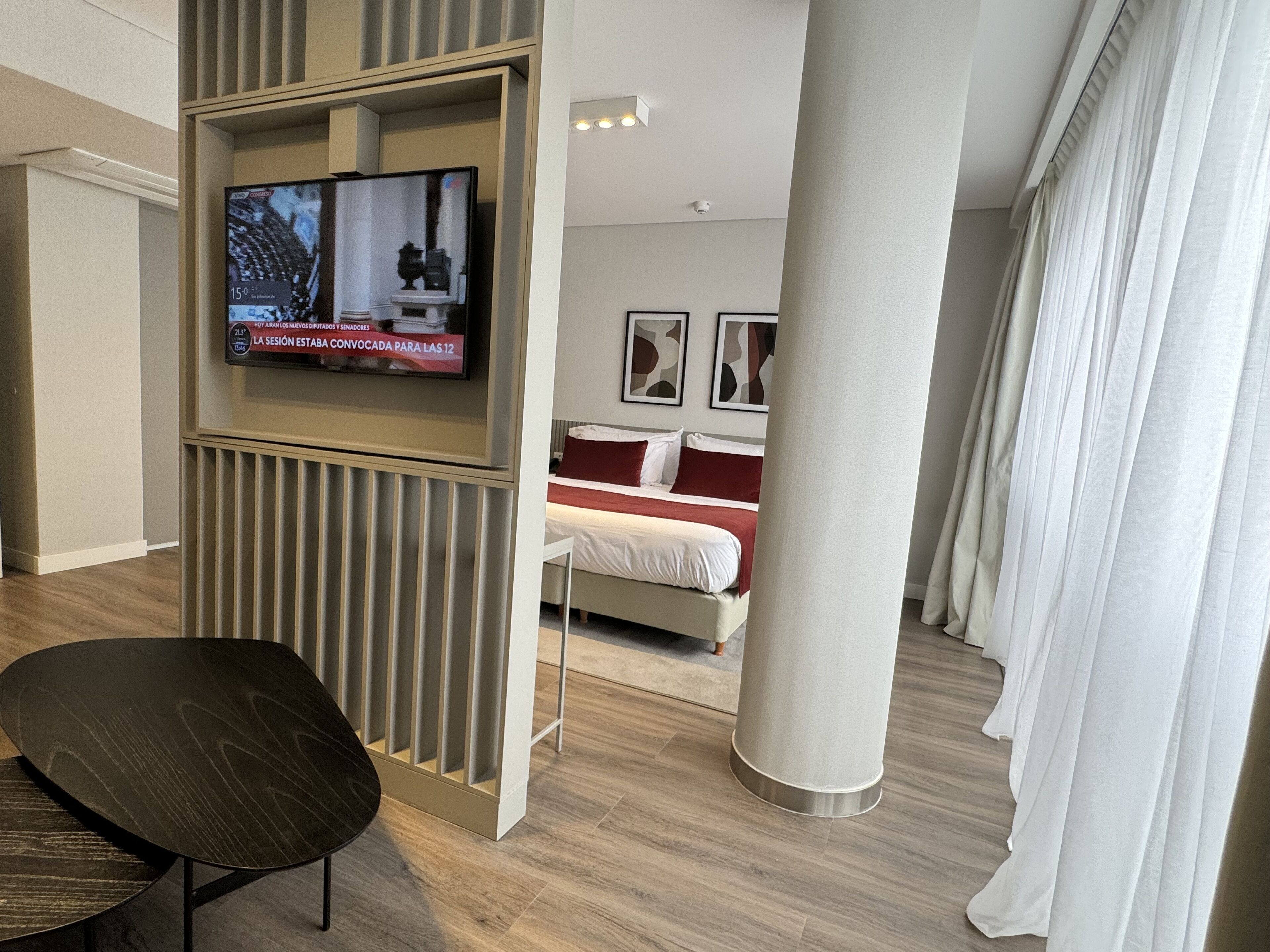 A hotel room with a TV mounted on a wall, a bed with red and white bedding, a wooden floor, and a large window with white curtains.