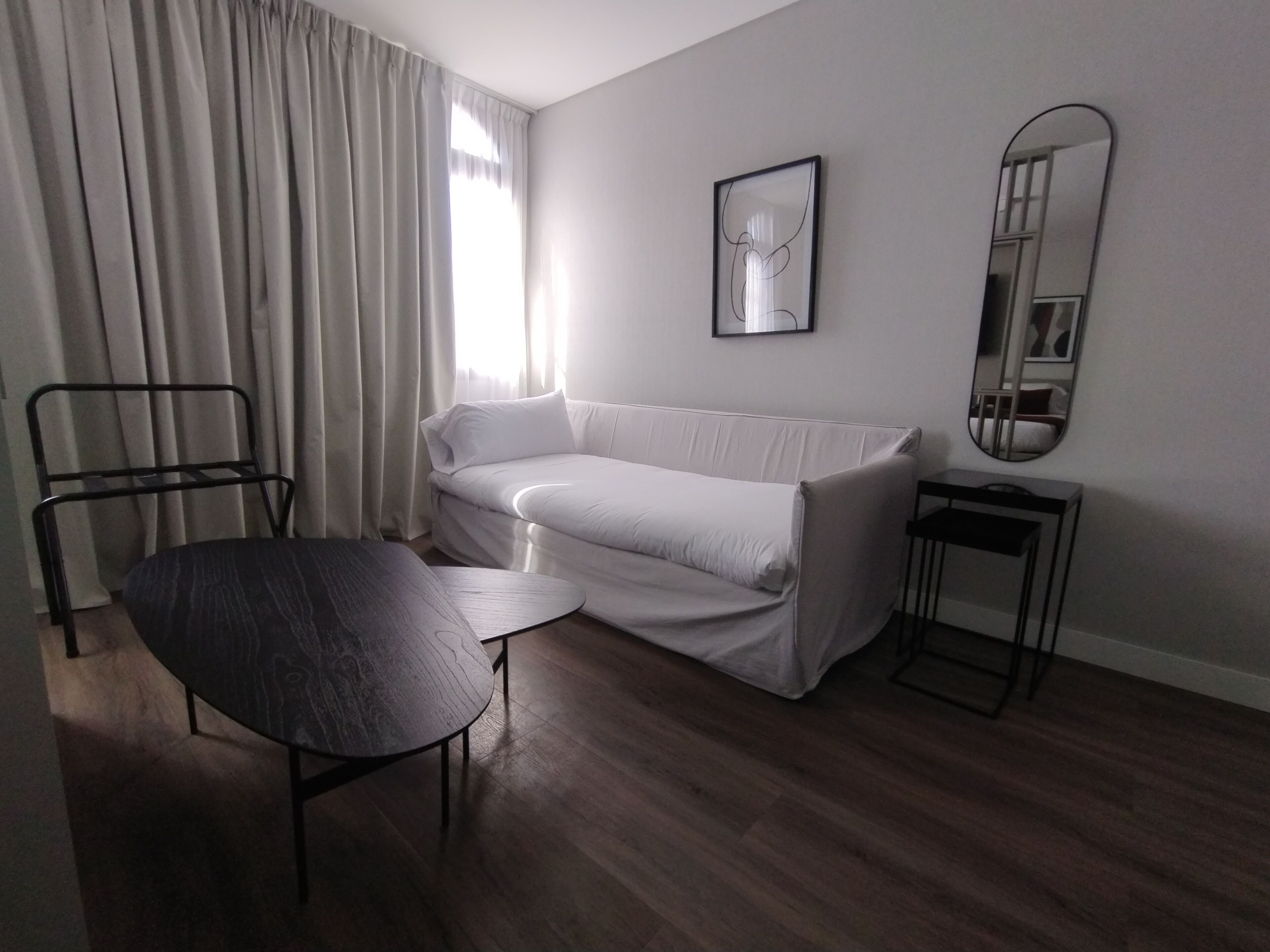 Habitació moderna i minimalista amb un sofà blanc, una taula rodona petita, una cadira de metall negre i un mirall sobre una taula auxiliar.