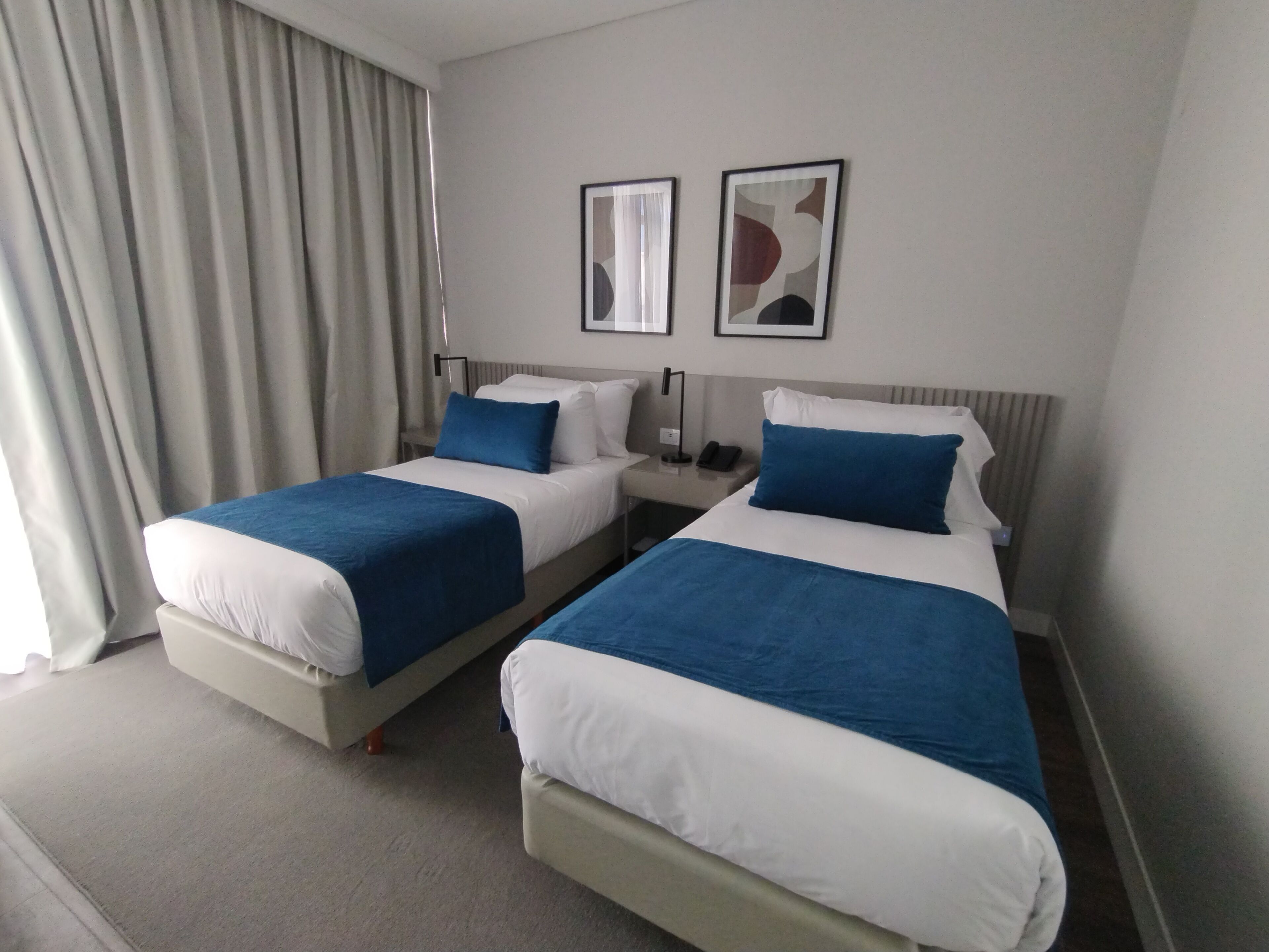A hotel room with two beds, each with blue and white bedding, a nightstand with a phone, and two framed pictures on the wall.