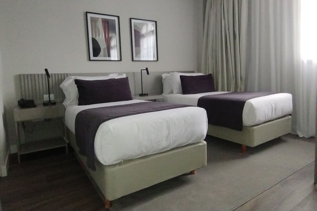 Ett hotellrum med två sängar, ett nattduksbord, en lampa och ett fönster med gardiner.