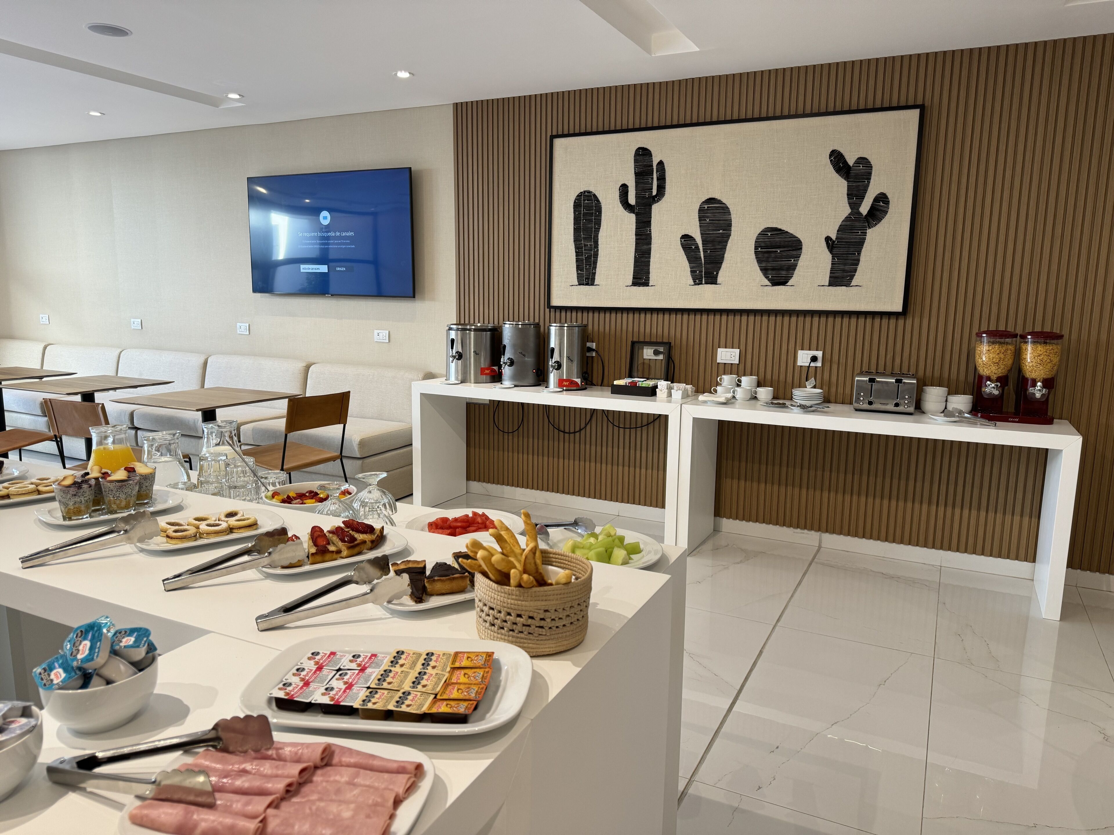 Une chambre d’hôtel moderne dotée d’un buffet de petit-déjeuner, d’une télévision à écran plat et d’une œuvre d’art sur le thème des cactus au mur.
