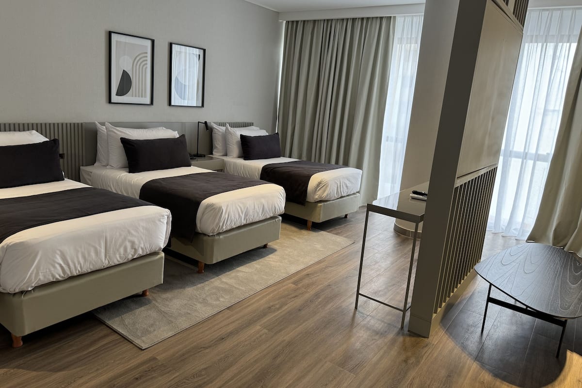 Ett hotellrum med tre sängar, ett skrivbord och ett stort fönster med gardiner.
