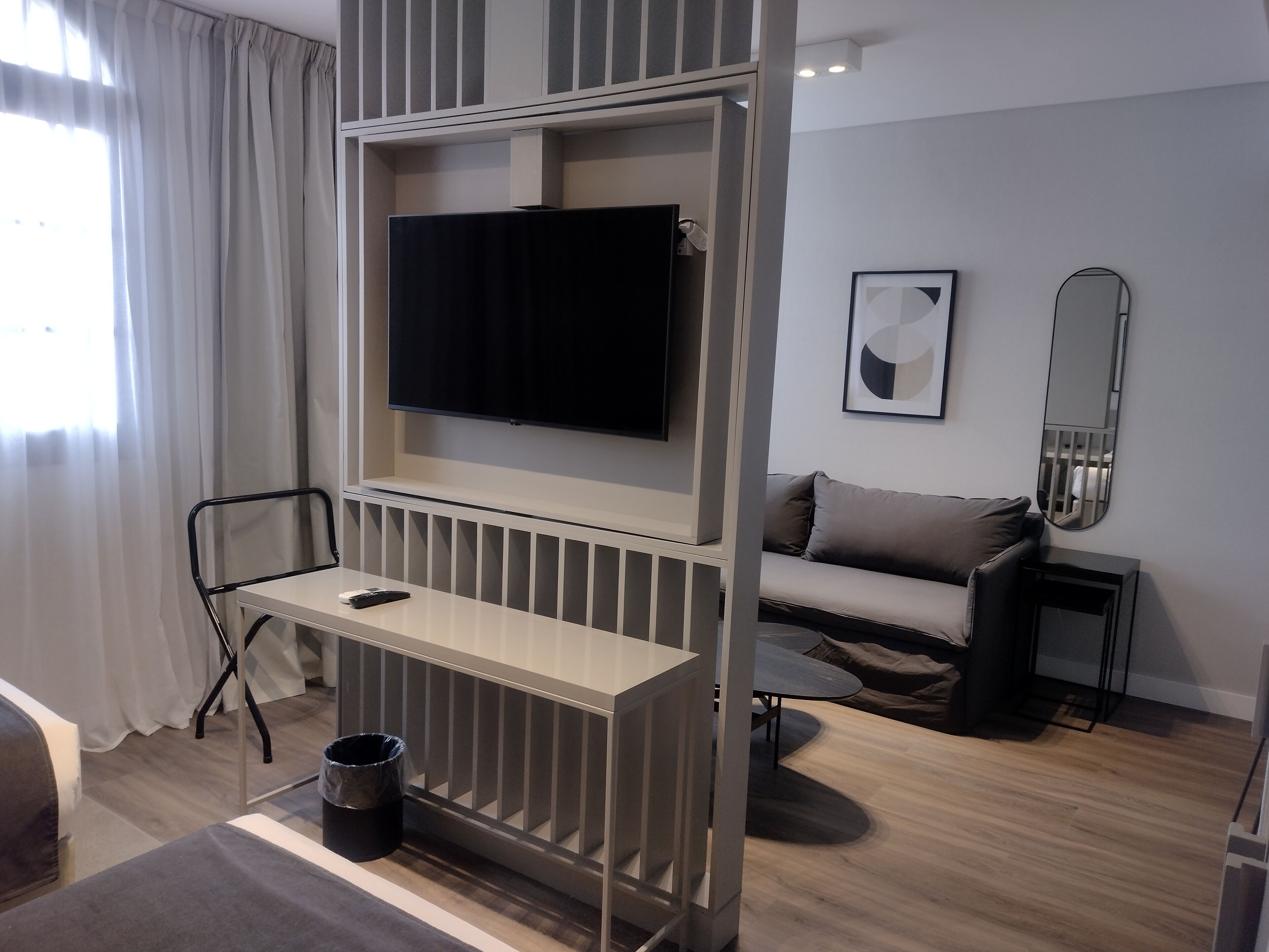 A modern hotel room with a TV mounted on a wall, a small table, a sofa, a mirror, and a trash bin.