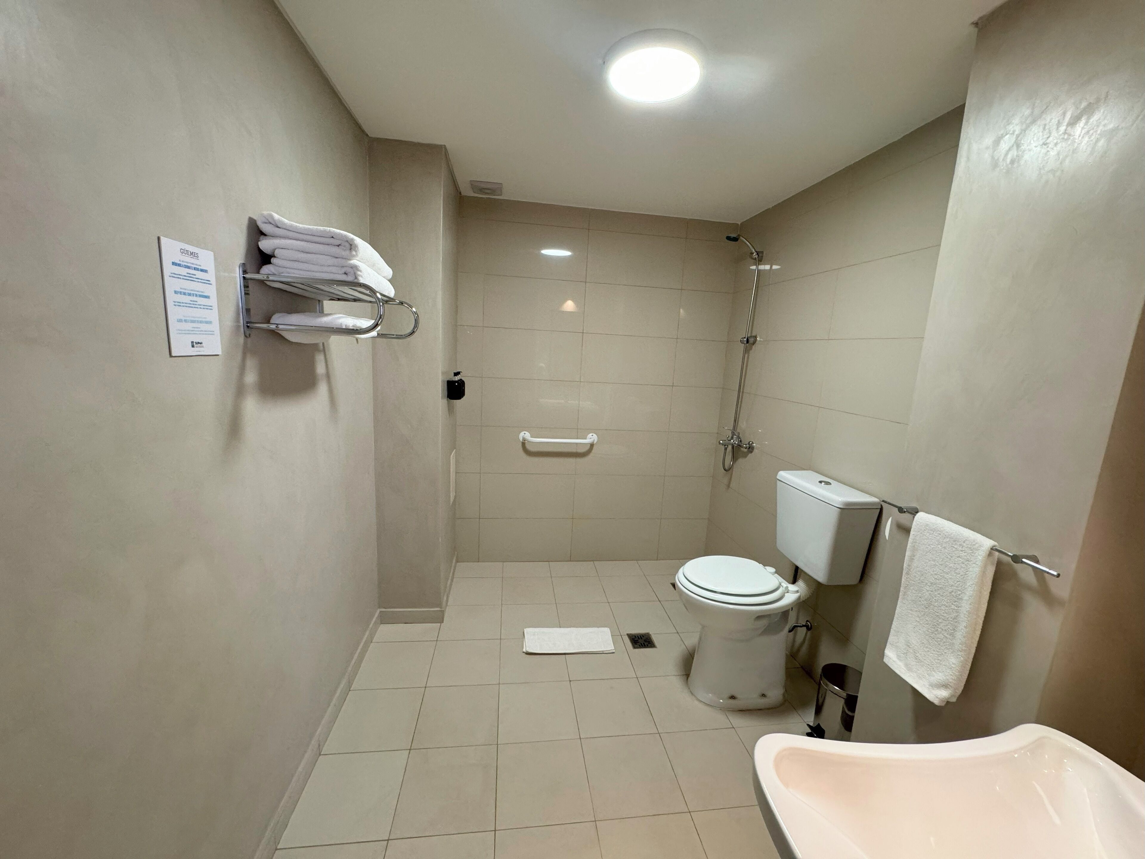 A small, clean bathroom with a toilet, sink, and shower. There are towels on a rack and a sign on the wall.