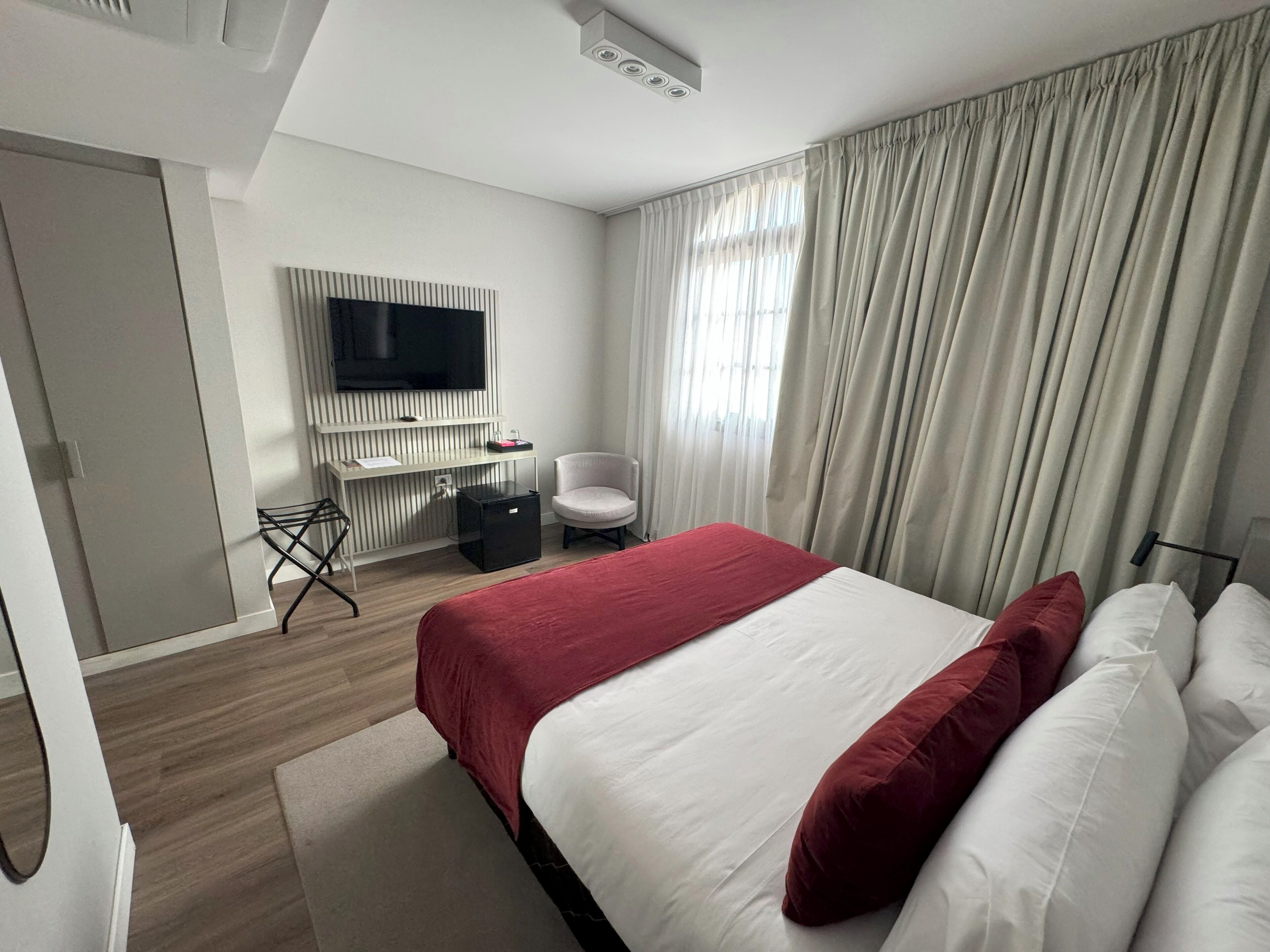 Una habitación de hotel moderna con una cama grande, un televisor de pantalla plana en la pared, un escritorio con una silla y una ventana con cortinas.