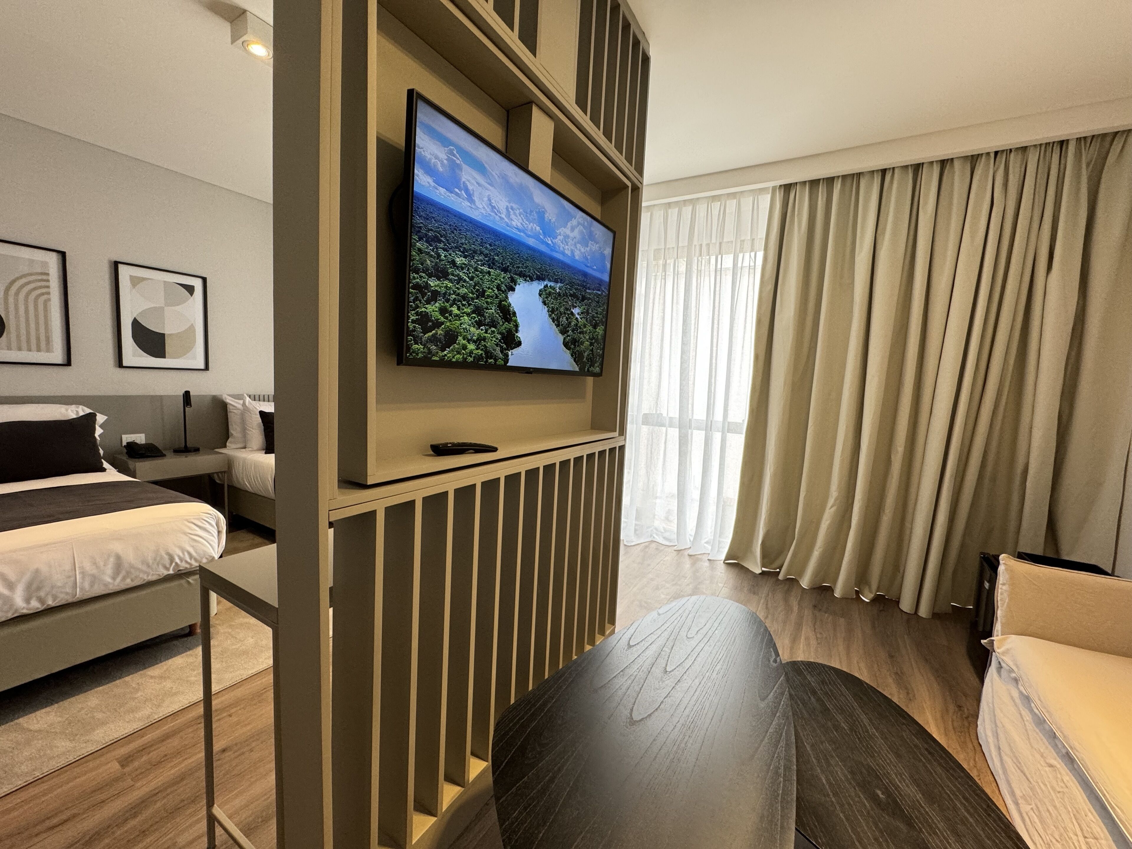 A modern hotel room with a large flat-screen TV mounted on a wall, a bed with a headboard, a seating area with a sofa and a table, and a window with curtains.