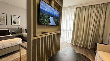 Una habitación de hotel moderna con una televisión de pantalla plana grande montada en la pared, una cama con cabecero, una zona de estar con un sofá y una mesa, y una ventana con cortinas.