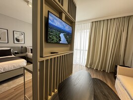 Una habitación de hotel moderna con una televisión de pantalla plana grande montada en la pared, una cama con cabecero, una zona de estar con un sofá y una mesa, y una ventana con cortinas.