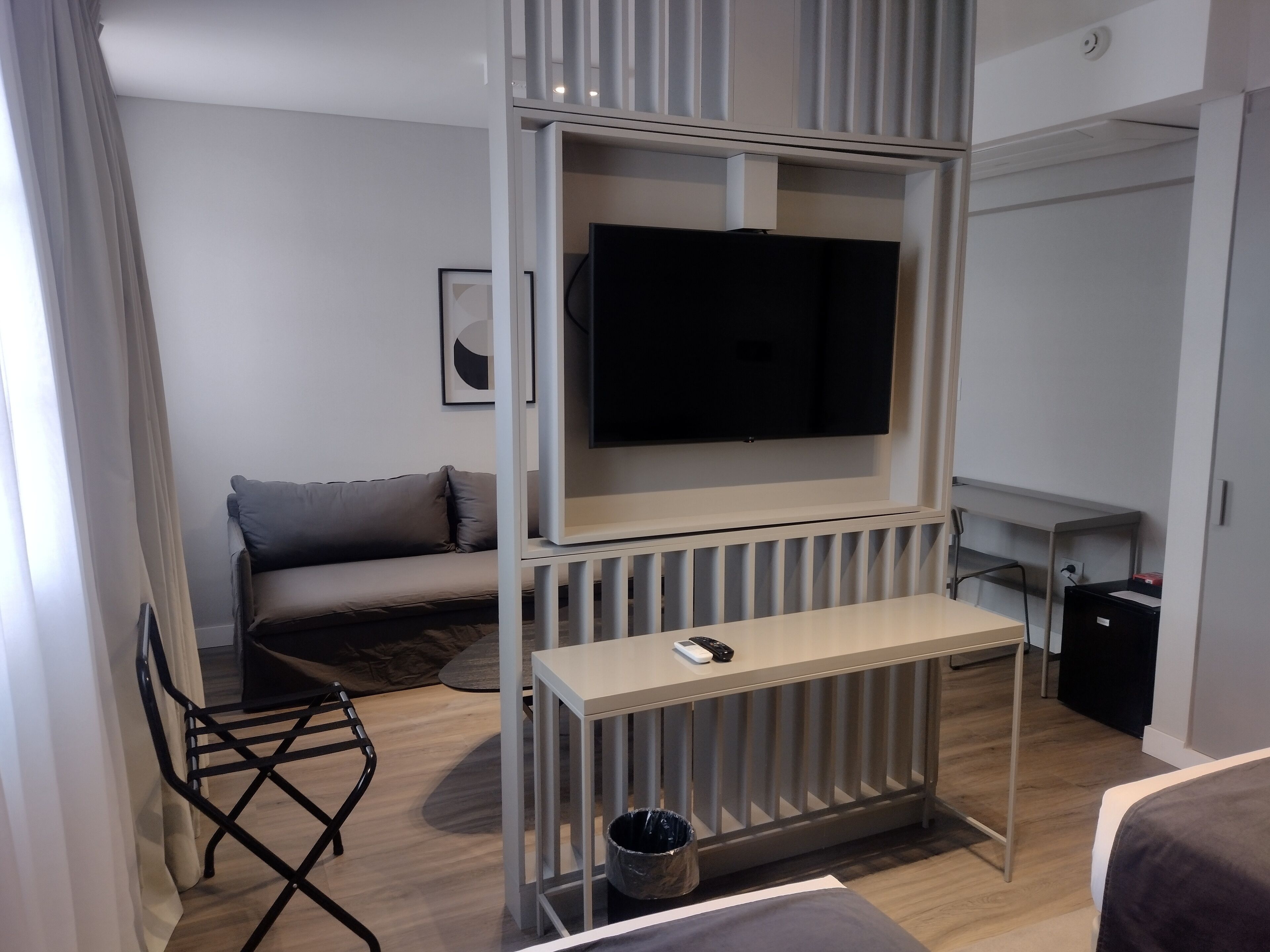 A modern hotel room with a grey sofa, a TV mounted on a white wall, a small table with a remote control, and a black trash bin.