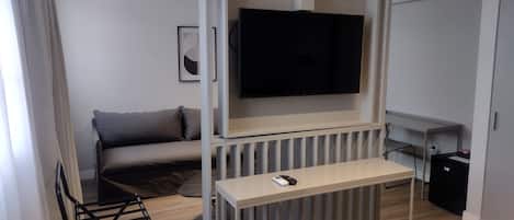 Une chambre d’hôtel moderne avec un canapé gris, un téléviseur fixé à un mur blanc, une petite table avec une télécommande et une poubelle noire.