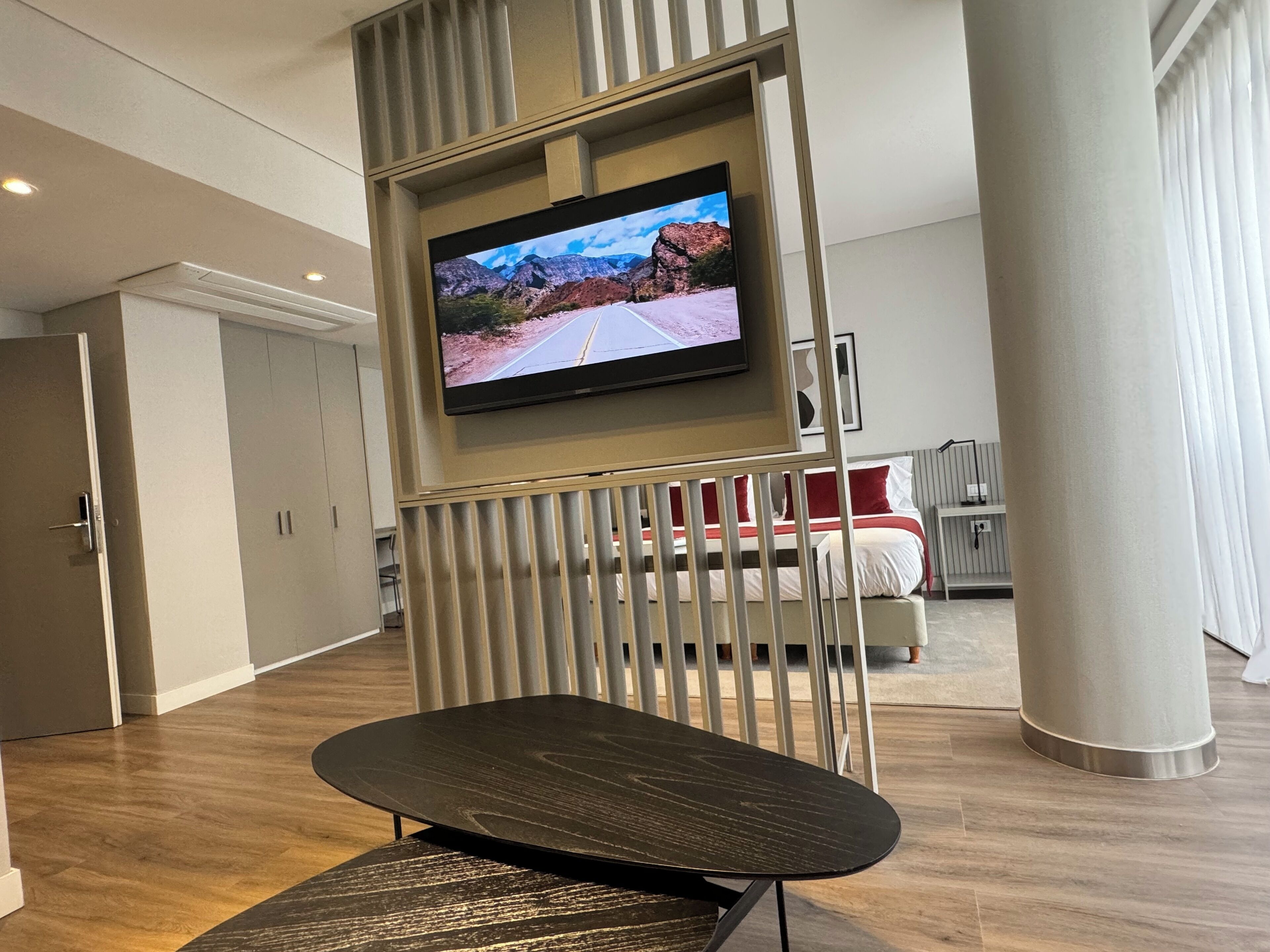 Una habitación de hotel moderna con una televisión de pantalla plana en la pared, piso de madera, cama con ropa de cama roja y blanca, y un pilar grande.