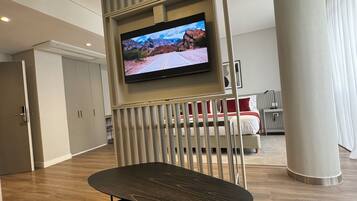 A modern hotel room with a flat-screen TV mounted on a wall, a wooden floor, a bed with red and white bedding, and a large pillar.