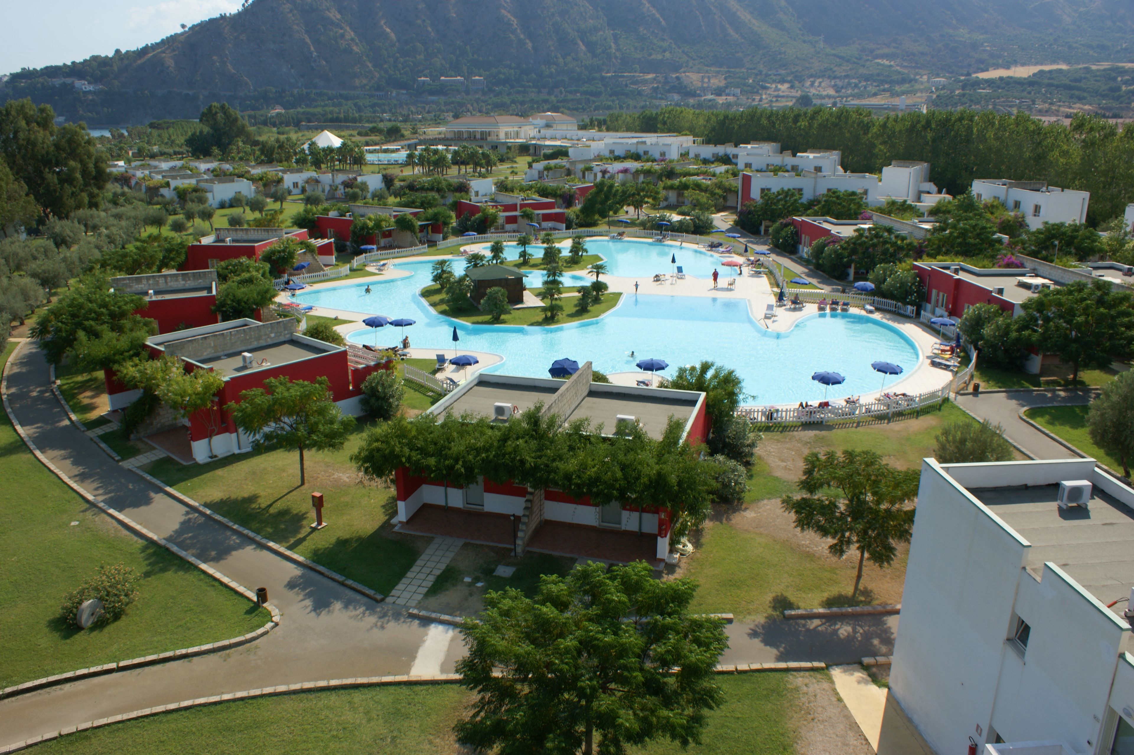 A resort with a large central swimming pool, surrounded by guest villas and lush greenery.